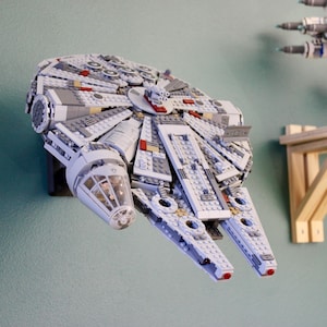 Puede incluir: Un modelo detallado de la nave espacial Millennium Falcon, construido con bloques de construcción entrelazados, está montado en una pared. El modelo es principalmente gris con detalles en rojo y blanco. Otro modelo de nave espacial es visible en una estantería en el fondo.