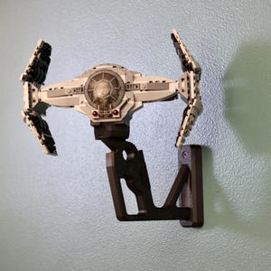 以下が含まれることがあります： 白と黒のTIEファイターモデルとダークブラウンの壁掛け。TIEファイターは壁に翼を広げて取り付けられています。