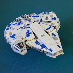 Puede incluir: Modelo Lego blanco y azul de la nave espacial Halcón Milenario. El modelo presenta una construcción detallada con varios paneles y una cabina. La nave se encuentra sobre un fondo verde azulado, mostrando su diseño icónico.
