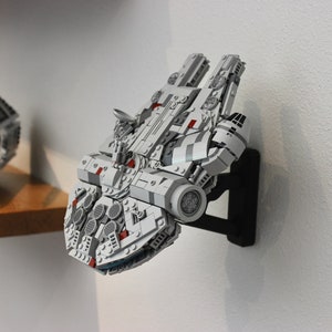 May include: A white and grey Lego Millennium Falcon spaceship model mounted on a black wall mount. The model has red details and is displayed on a white wall.