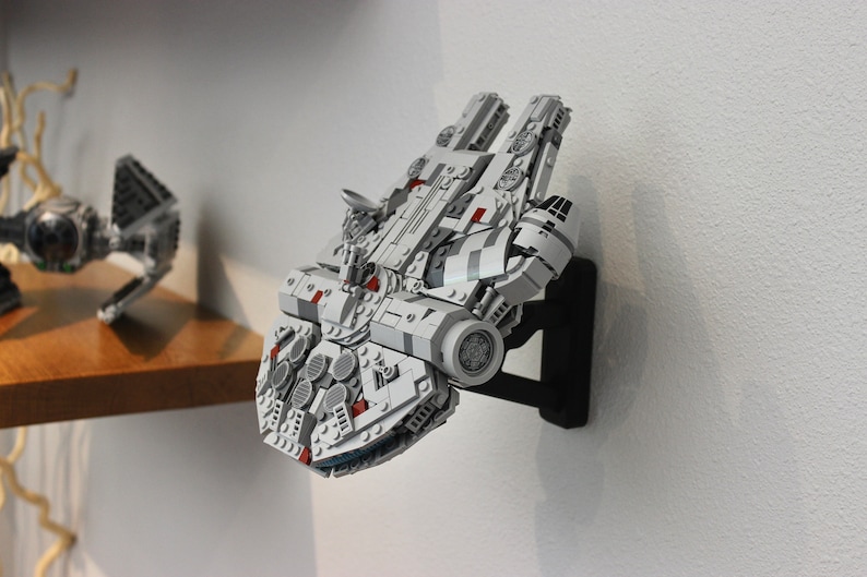 May include: A white and grey Lego Millennium Falcon spaceship model mounted on a black wall mount. The model has red details and is displayed on a white wall.