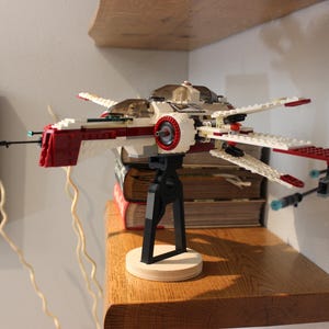 May include: A red and white Lego spaceship model with a black stand. The spaceship has a red cockpit and white wings. The stand is made of black plastic and has a round base.
