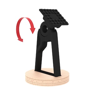 Puede incluir: Un soporte de teléfono negro con una parte superior giratoria y una base de madera. El soporte está diseñado para sostener un teléfono en posición vertical.