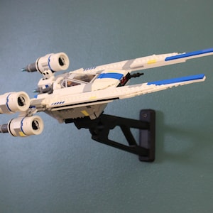 Puede incluir: Un modelo blanco y azul de un caza estelar X-wing, montado en un soporte de pared negro. El modelo presenta componentes detallados del motor y detalles azules en las alas. El caza estelar es un artículo popular en la ciencia ficción.