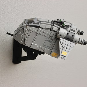 Puede incluir: Una nave espacial Lego gris y plateada con una cabina transparente y dos puertos de escape negros. La nave espacial está montada en un soporte de pared negro.