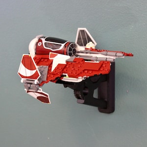 以下が含まれることがあります： 黒色の壁掛けに、赤と白のレゴスターウォーズV-4X-Dスターファイターモデルが取り付けられています。モデルは白いコックピットと灰色の翼を持っています。