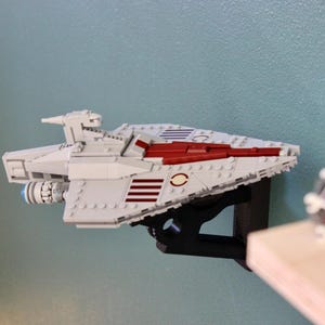 Op de afbeelding: Een wit en rood Lego-ruimteschipmodel gemonteerd op een zwarte wandbevestiging. Het ruimteschip heeft een rode streep en een witte streep aan elke kant.