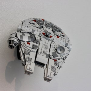 May include: A white and grey Lego model of the Millennium Falcon spaceship from Star Wars. The model has red accents and is mounted on a white wall.