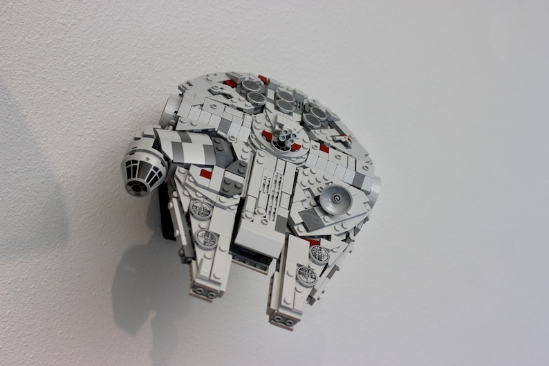 May include: A white and grey Lego model of the Millennium Falcon spaceship from Star Wars. The model has red accents and is mounted on a white wall.