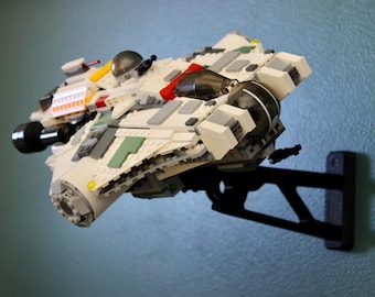 May include: A white, gray, and green Lego spaceship model mounted on a black wall bracket. The model features various details, including red and yellow accents, and a black cylindrical engine. The spaceship is angled, giving a sense of flight.