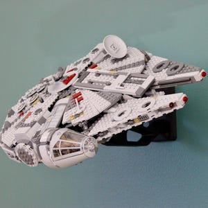 Könnte beinhalten: Ein detailliertes Modell des Raumschiffs Millennium Falcon, aus ineinandergreifenden Kunststoffbausteinen. Das Modell ist überwiegend weiß und grau, mit roten und schwarzen Akzenten. Es ist auf einem schwarzen Präsentationsständer an einer hellblauen Wand montiert.
