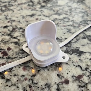 May include: A white LED light fixture with a clear, circular lens. The fixture is mounted on a white base with two mounting holes and a white wire. The light is on, emitting a warm, yellow glow.