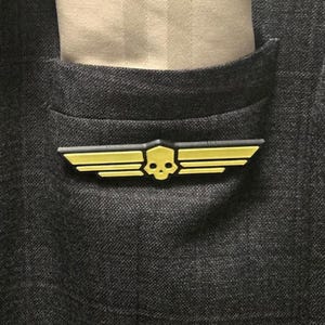 May include: A yellow and black pin featuring a stylized skull centered between two sets of horizontal wings. The pin is attached to the front of a dark gray textured fabric pocket, with a light beige fabric visible inside the pocket opening.