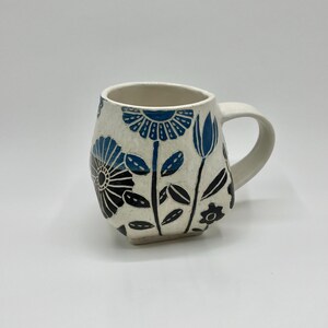 May include: Cream-colored ceramic mug with a floral design in black and blue. The flowers and leaves are hand-painted on the mug's surface. The mug has a rounded shape and a curved handle.