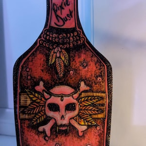 May include: A wooden bottle shaped cutting board with a red, orange, and yellow skull and crossbones design. The bottle is labeled "Pixie Dust" and has a cork on top.