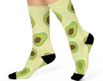May include: A pair of cream-colored socks with a repeating pattern of green avocado halves. The socks have black heels and toes.