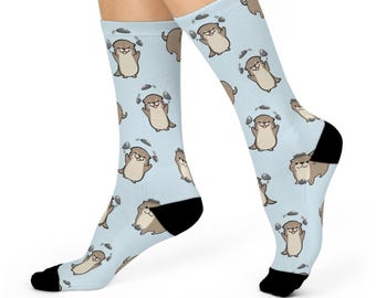 May include: A pair of light blue socks with a repeating pattern of brown otters juggling gray rocks. The socks are shown on a model's feet.