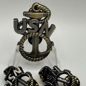 May include: Three gold-toned metal US Navy anchor pins. The largest pin is on a display stand. The smaller pins have a gold-toned finish with a black backing.