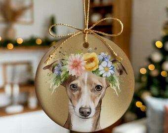 May include: A round, gold-toned ceramic ornament featuring a detailed illustration of a brown and white dog wearing a colorful floral crown of pink, yellow, and blue flowers. The ornament is suspended by a thin gold string tied in a decorative bow.