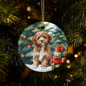 May include: Round Christmas ornament featuring a watercolor painting of a brown and white puppy sitting in front of a Christmas tree with red ornaments and wrapped gifts. The ornament has a gold string for hanging.