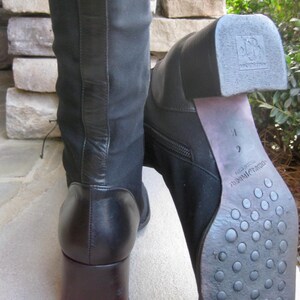 Puede incluir: Dos botas negras hasta la rodilla con tacones cuadrados marrones oscuros. Una bota est&aacute; de pie, la otra est&aacute; inclinada, mostrando la suela con un patr&oacute;n texturizado. Las botas tienen una cremallera lateral y un panel de cuero en la parte trasera.