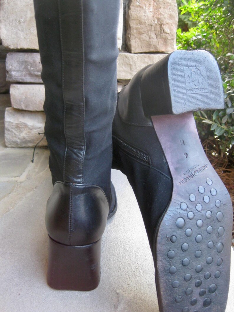 Puede incluir: Dos botas negras hasta la rodilla con tacones cuadrados marrones oscuros. Una bota est&aacute; de pie, la otra est&aacute; inclinada, mostrando la suela con un patr&oacute;n texturizado. Las botas tienen una cremallera lateral y un panel de cuero en la parte trasera.