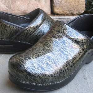 May include: Pair of black clogs with a glossy finish and a unique gold and white scribble pattern. The shoes have a rounded toe and a thick sole. The clogs are made of a durable material and are suitable for various activities.
