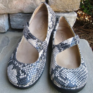 May include: A pair of black and white snake-skin patterned shoes with a strap across the top. The shoes have the word "Vionic" written on the inside of each shoe.