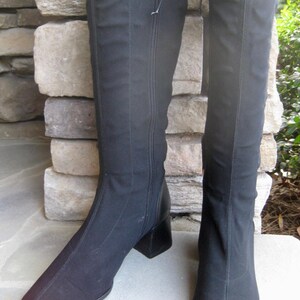 Puede incluir: Un par de botas negras hasta la rodilla con punta cuadrada y tac&oacute;n de bloque. Las botas tienen cierre de cremallera lateral y est&aacute;n hechas de tejido el&aacute;stico. Las botas se exhiben sobre una superficie de piedra.