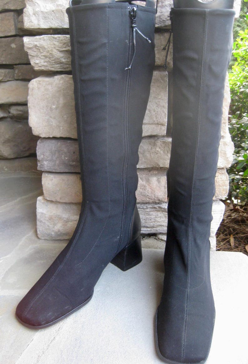 Puede incluir: Un par de botas negras hasta la rodilla con punta cuadrada y tac&oacute;n de bloque. Las botas tienen cierre de cremallera lateral y est&aacute;n hechas de tejido el&aacute;stico. Las botas se exhiben sobre una superficie de piedra.