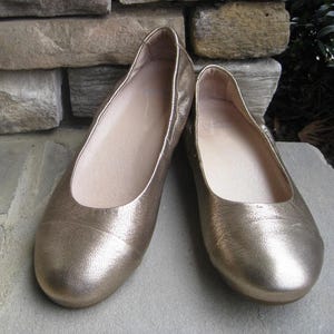 May include: A pair of gold-colored ballet flats. The shoes have a rounded toe and a low heel. The interior of the shoe is a light beige color. The shoes are made of a textured material.