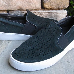 May include: A pair of dark green slip-on sneakers with white soles. The uppers feature a floral cut-out design. The shoes have a rounded toe and a black elastic band across the top.