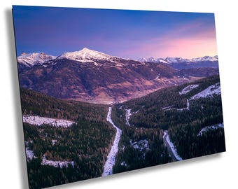 May include: An aerial view of a snow-capped mountain range with a winding road leading through a forest. The sky is a vibrant purple and pink, creating a beautiful sunset scene.