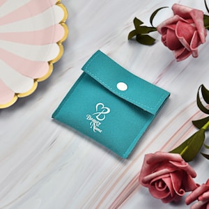 Może przedstawiać: Torebka na biżuterię z turkusowego aksamitu z białym zapięciem na guzik. Torebka ma białe logo z napisem "Beauty Name" ze stylizowanym sercem i literą B.