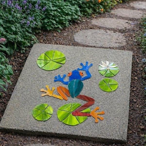 May include: A square concrete garden stepping stone featuring a mosaic design of a frog in blue, orange, and red glass pieces, surrounded by several green glass lily pads and a single white and green glass water lily flower embedded in the surface.