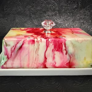 May include: A rectangular butter dish featuring a white base and a lid decorated with a vibrant, abstract alcohol ink pattern in shades of hot pink, yellow, and green. The lid is topped with a clear, faceted, diamond-shaped glass knob.