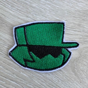 May include: An embroidered patch in green and black, with a white border. The patch depicts a stylized hat and sunglasses design. Suitable for application on clothing or accessories.