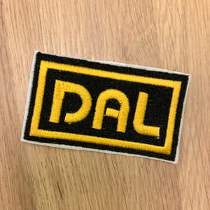 May include: Embroidered patch featuring the letters "DAL" in yellow, set against a black background with a yellow border. The patch has a white border and is on a wooden surface.