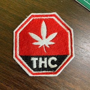 May include: Embroidered patch in a stop sign shape with a red background and a white cannabis leaf. The bottom section is black with the letters "THC" in white. The patch has a white border.