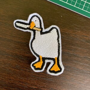 Peut inclure: Écusson brodé d'un canard blanc avec un contour noir, un bec et des pattes orange. Le canard tient un objet marron dans son bec. L'écusson a une bordure blanche et est sur une surface en bois foncé.