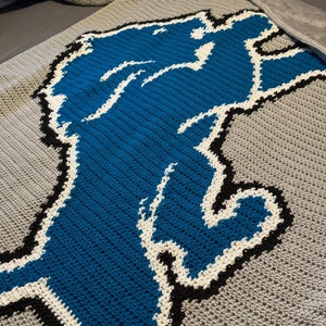 May include: A crocheted blanket with a blue and white lion design on a gray background.