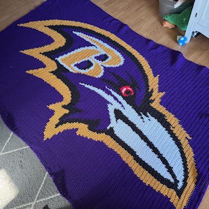 May include: A purple knitted blanket featuring the Baltimore Ravens logo. The logo is a stylized raven's head with gold, black, light blue, and red accents. The blanket is made of a soft, textured material.