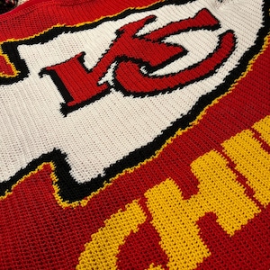 Op de afbeelding: Gehaakte deken met het logo van de Kansas City Chiefs in rood, wit, zwart en geel.