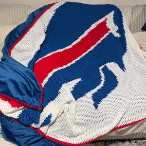 Op de afbeelding: Een gehaakt deken met het logo van de Buffalo Bills in blauw, rood en wit. Het deken is gevouwen en over een bank gelegd.
