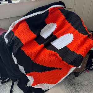 Könnte beinhalten: Eine gehäkelte Decke mit einem schwarz-weiß-orangenen Streifenmuster. Die Decke ist in der Mitte gefaltet und hat ein weißes Häkelmuster.