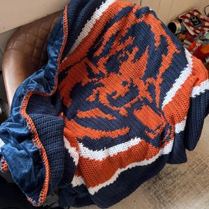 May include: A crocheted blanket featuring a tiger face design in orange, white and navy blue. The blanket is folded and draped over a brown leather chair.