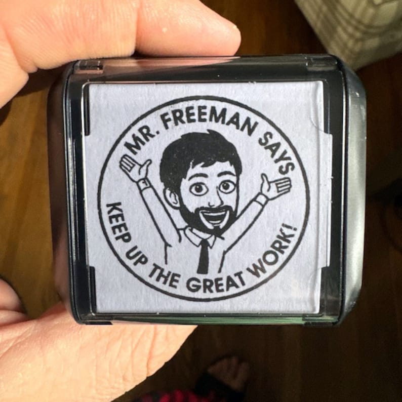 May include: A black and clear stamp with the text "MR. FREEMAN SAYS" and "KEEP UP THE GREAT WORK!" surrounding a cartoon image of a man with his arms raised. The stamp is held by a hand.