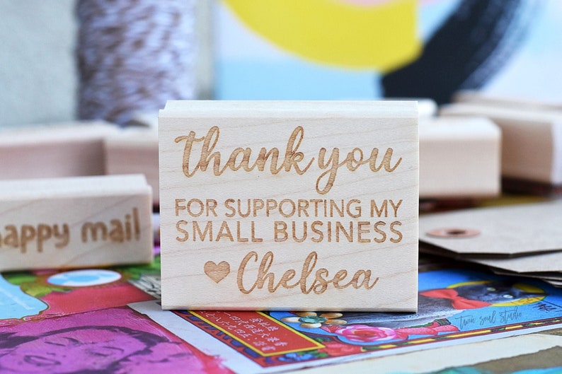 Pu&ograve; includere: Un timbro in legno con le parole "thank you for supporting my small business Chelsea" incise in un carattere corsivo. &Egrave; incluso un simbolo a forma di cuore. Altri timbri e articoli di cancelleria sono sullo sfondo.