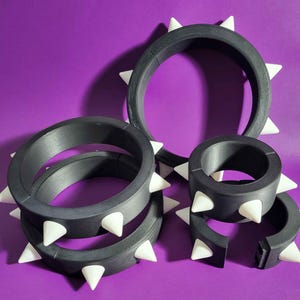 May include: A collection of black, spiked bracelets and cuffs with white conical spikes. The jewelry is arranged on a purple background. The bracelets and cuffs vary in size and shape, with some being full circles and others open.