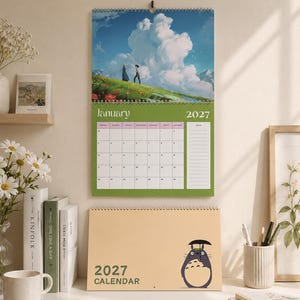 May include: A 2027 wall calendar featuring a top section with an illustration of two figures on a grassy hill under a large cloud and a bottom section displaying the month of January 2027. Below, a desk calendar shows a cartoon creature with an umbrella.